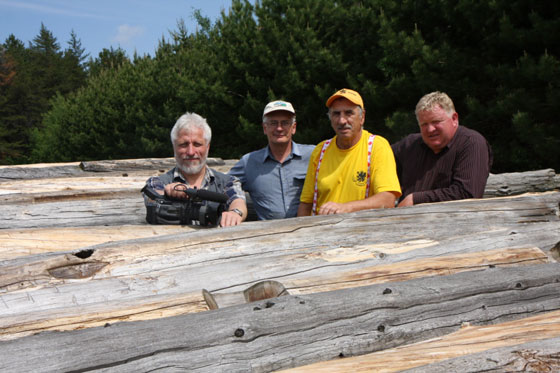 Four men stand among round logs